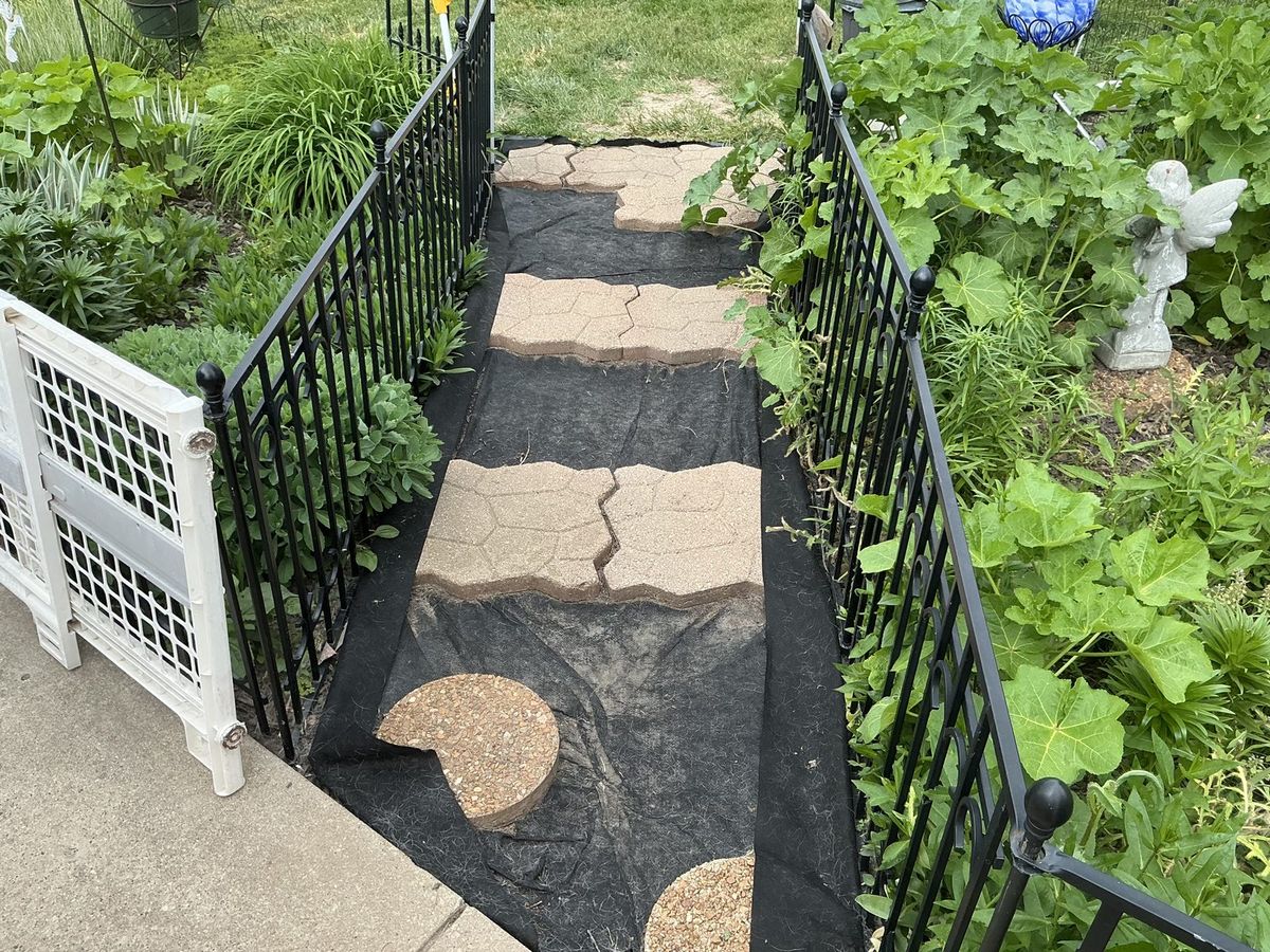 Stepping-stone path on landscape fabric between garden beds and black fencing, before